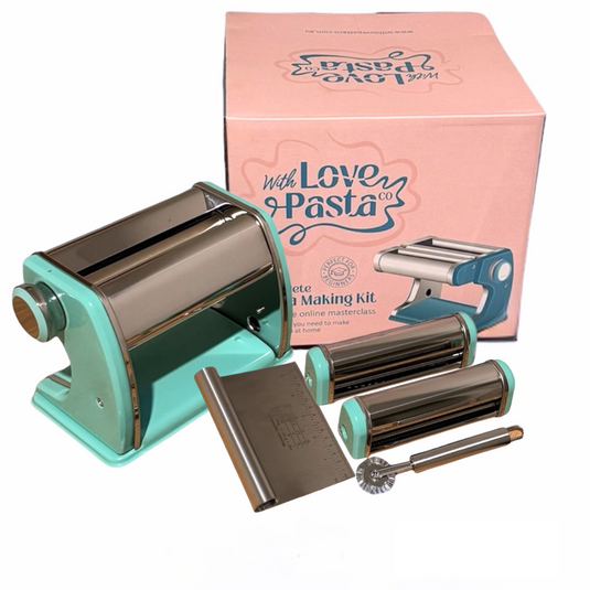 A stainless steel pasta making kit with teal accents, including a pasta machine, spaghetti and fettuccine cutters, crank handle, cleaning brush, ravioli cutter wheel, and dough scraper, presented next to its pink packaging box.